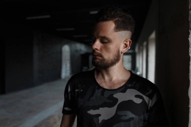 Yakışıklı genç adam şık saç modeli ve bir modern moda askeri t-shirt bir karanlıkta kapalı poz içinde acımasız sakal. Çekici adam