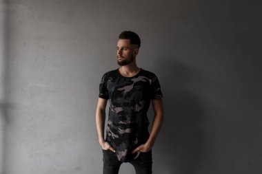 Yakışıklı genç adam şık saç modeli ve bir askeri kamuflaj moda t-shirt ayakta gri bir arka plan karşı sokakta acımasız sakal. Moda çekici modern adam