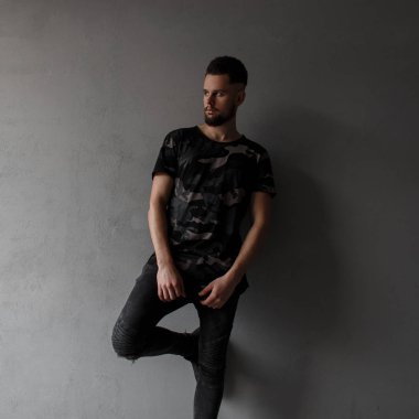 Hipster çekici genç adam şık saç modeli ve acımasız sakal bir askeri moda gömlek, siyah kot ve gri bir duvar yakınındaki popüler spor ayakkabı modeli. Şık modern adam