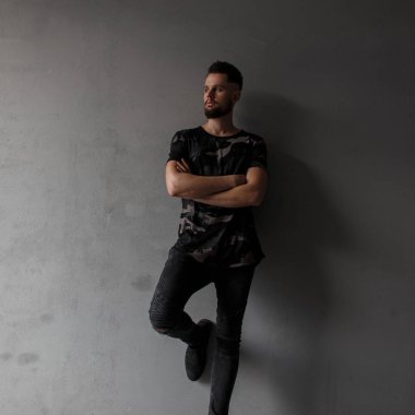 Gri bir duvara poz hipster çekici genç adamla bir moda saç modeli ve askeri bir şık gömlek, siyah kot ve trendy spor ayakkabı, acımasız sakal. Şık modern moda adam