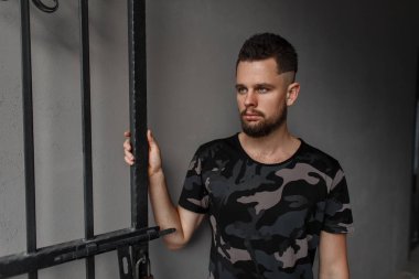 Bir saç modeli ve gri arka plan üzerinde metal kapılar yakınındaki bir moda askeri yeşil kamuflaj tişört ayakta şık sakal oldukça yakışıklı hipster genç adamla. Moda çekici adam