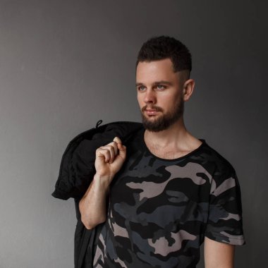 Acımasız genç hippi erkek saç modeli ve şık sakal moda askeri gri kamuflaj tişört ve siyah sonbahar ceket ile sokakta gri duvarının yakınında duruyor. Moda çekici adam