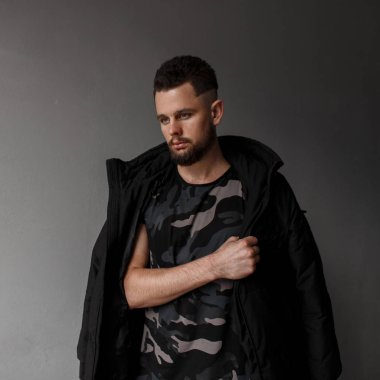 Yakışıklı genç adam saç modeli ve şık sakallı bir moda askeri yeşil kamuflaj tişört ve siyah sonbahar ceket poz gri bir arka plan yakın. Moda çekici adam
