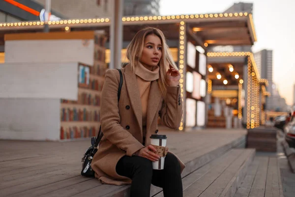 Örme vintage bej kazak, kot pantolon ve şık bir siyah çanta şık bir ceket genç kadın bir ahşap merdiven üzerinde oturur ve sıcak tatlı kahve sokakta tutar. Çekici kız