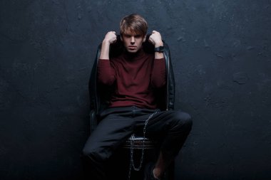 Genç hipster adam şık vintage ceket siyah spor ayakkabı gümüş metal zinciri içinde şık Jeans kırmızı Golf oturur ve bir odada duvara karşı sandalyeye teşkil etmektedir. Çekici adam