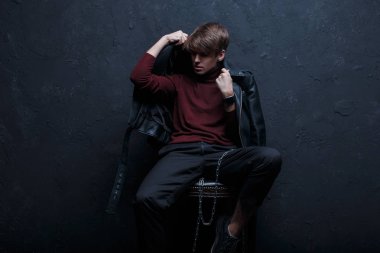 Bir metal zincir ile siyah spor ayakkabı içinde şık Jeans kırmızı Golf şık vintage ceketli genç hipster adamı oturur ve bir sandalyeye duvarının yakınında bir stüdyoda teşkil etmektedir. Çekici trendy modern adam