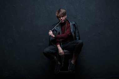Vintage ceket kot pantolon ve spor ayakkabı Bordo Golf bir saç modeli genç seksi adamla bir sandalyeye oturur ve duvarın yanında karanlık bir stüdyoda metal bir zincir Dişlerini içinde tutar. Modern adam. Moda