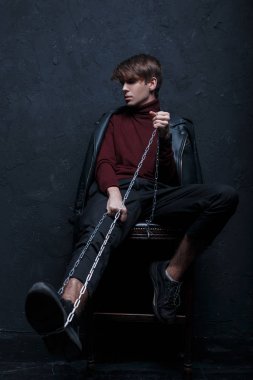 Genç modern insan olarak vintage Bordo golf modaya uygun kot pantolon ve bir metal zincir ile siyah spor ayakkabı moda ceket siyah duvarının yakınında bir odada bir tahta sandalyede oturur. Yakışıklı olan şık