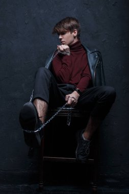 Genç bir adam siyah deri vintage ceket siyah pantolon ve şık spor ayakkabıları modaya uygun bir Bordo Golf bir tahta sandalyede oturur ve bir metal zincir Studio duvarın yanında tutar. Şık adam