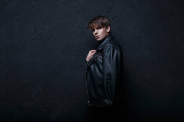 Siyah duvarın yanında karanlık bir odada kamera bakarak ve ayakta siyah kot ile retro tarzında bir siyah deri ceketli bir saç modeli olan moda serin genç adam. Güzel şık adam. 