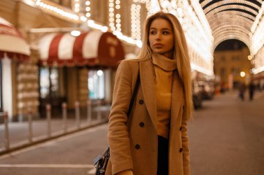 Güzel genç kadın şık bej bir palto siyah şık kot pantolon ve siyah deri çanta içinde bir örme vintage süveterli şehrin içinden Geceleri ışıkları bir arka plan üzerinde yürür. Sevimli kız