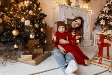 Vintage kırmızı kazaklı genç neşeli kadın anne stüdyo ahşap zemin üzerinde oturur ve onun kollarında bir çocuk şık Noel peluş giysileri beyaz bir şömine ve Noel ışıkları yakın tutar.