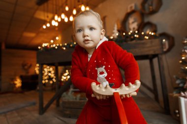 Sevimli tatlı küçük kız bebek Festival çelenk ve ışıklar bir stüdyo ahşap kızıl geyik sandalyede oturan kırmızı peluş şık takım elbise dekore edilmiş bir Noel. Yeni yıl atmosfer. Noel ruh hali