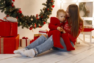 Genç kadın anne örme kırmızı kazaklı stüdyoda bir ahşap zemin üzerinde oturur ve arka planı Noel süslemeleri yılbaşı peluş giysili bir çocuk öpücük. Hafta sonu aile ile. 