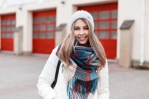一个开朗积极的年轻女子的肖像与甜蜜的微笑与温暖时尚格子围巾和白色时尚夹克的背景下 一个红色的门建筑 女孩笑了 — 图库照片