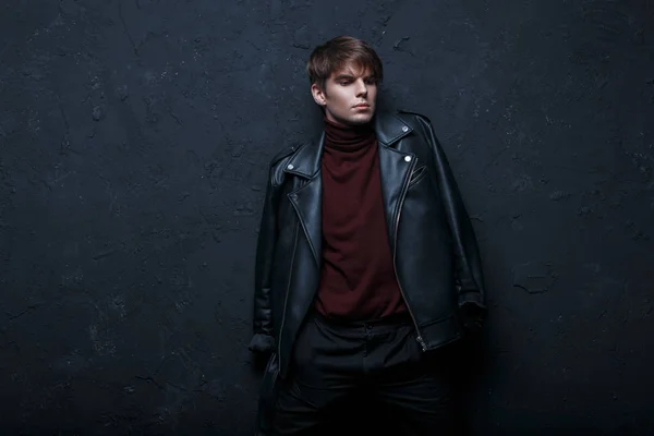 Attractive handsome young man in a black vintage autumn jacket in a stylish burgundy golf in black jeans is standing and posing indoors against a black wall. Stylish guy model
