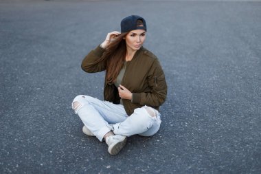 Güzel genç kadın moda basketbol beysbol şapkası içinde şık, şık modern yeşil askeri ceket kot ve asfalt yolda oturup beyaz spor ayakkabı sökük. Çekici kız.