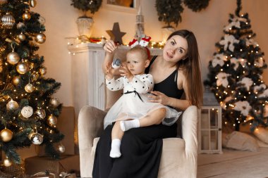 Genç kadın anne ile güzel bir güzel küçük kız şık elbiseler içinde sevimli oturma ve stüdyo Noel ağacı yakınındaki beyaz bir sandalyede andan zevk. Aile kutlama