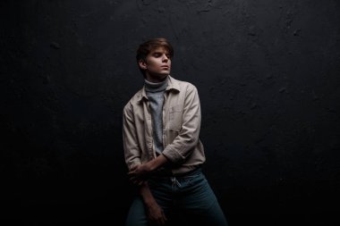 Şık güzel çekici genç adam şık vintage Mavi Jeans kapalı siyah bir duvara yakın poz gri süveterli moda ceket şık bir saç modeli ile. Amerikan modern adam modeli