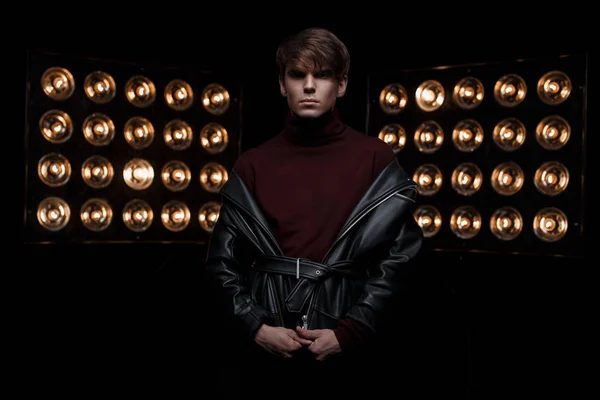 Attractive young stylish man in a leather jacket in burgundy stylish golf stands in a dark studio against the background of bright orange vintage paws of lights. Cute fashion star guy