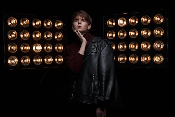 Amazing young man with a fashionable hairstyle in a black fashionable jacket in red stylish golf and black jeans is standing and posing in a dark room near bright vintage electric lamps. Cute guy
