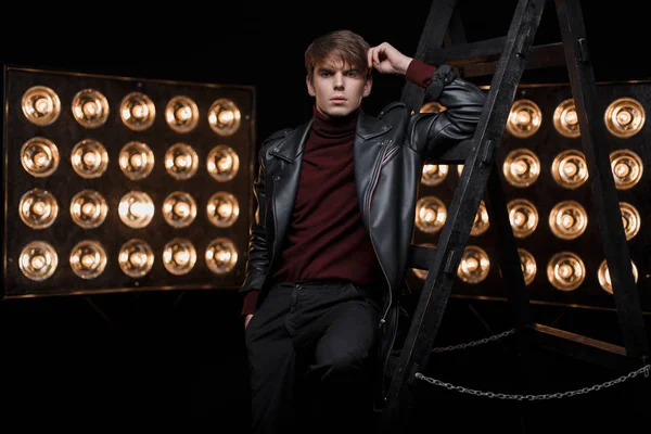 Fashionable young stylish guy in a red stylish golf in a vintage black jacket in black pants posing in the studio near the metal stairs against the background of bright yellow lamps.Fashionable guy