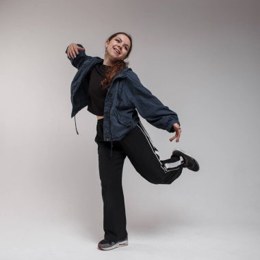 Neşeli genç kadın dansçı trendy siyah spor kahverengi uzun saçlı beyaz bir duvar yakınındaki bir stüdyoda kamera önünde hip hop dans hareketleri gösterir. Modern koreografi. Komik kız
