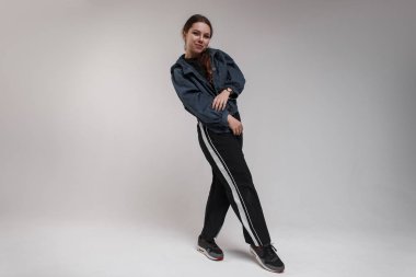 Şirin yetenekli genç kadın dansçı trendy spor giyim ve siyah spor ayakkabı beyaz bir duvar yakınındaki bir odada modern hip hop dans hareketleri gösterir. Tatlı hippi kız dans