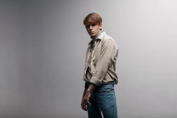 American young man with a fashionable hairstyle in a light, light, stylish jacket in retro style in blue vintage pants is standing in the studio near a white wall. Handsome guy. Modern youth style