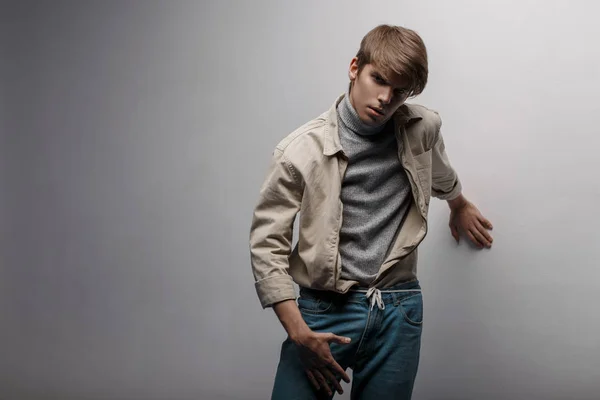 Young mysterious man with a fashionable hairstyle in a light, light jacket in knitted golf in blue fashionable pants is standing in a room near a white wall. American attractive fashionable guy.