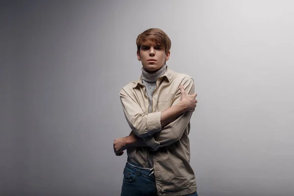 Serious young man with a fashionable hairstyle in a fashionable light jacket in gray golf in blue stylish pants posing in studio near a white wall. Stylish guy. Modern men\'s fashion