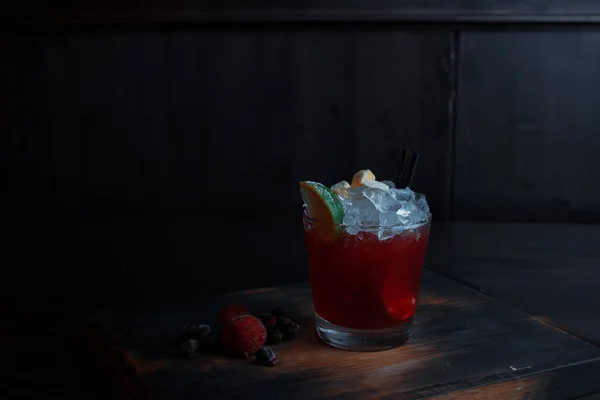 美味的甜红色酒精鸡尾酒与冰和樱桃糖浆与伏特加在水晶玻璃站在一个木桌上的酒吧在黑暗的背景 用新鲜浆果装饰的鸡尾酒 — 图库照片