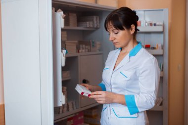 A young female doctor in white uniform is standing in a room for medical staff near a cabinet with pills and checks the shelf life of the drugs.