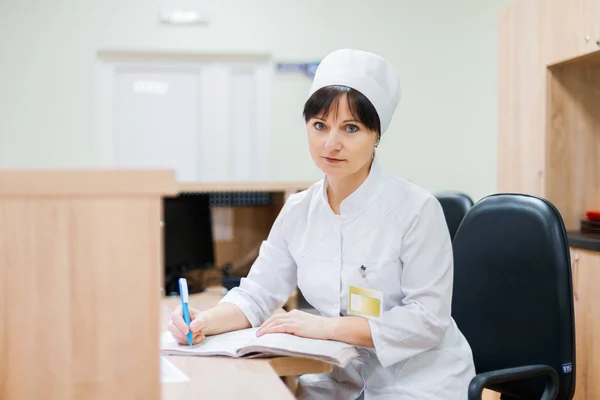 A female nurse in a white medical gown sits at a wooden desk at the reception desk and makes entries in a work log. Woman doctor working