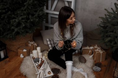 Siyah pantolon içinde şık bir vintage kazaklı genç çekici kadın Noel odası beyaz mumlar arasında ağacında yakın katta oturuyor ve hediyeler sevimli kız tatil hakkında düşünüyor.