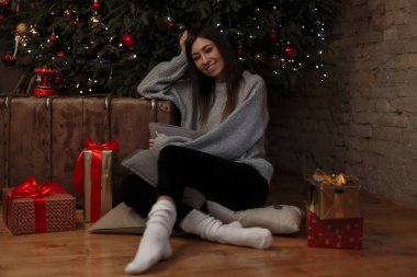 Sıcak örme kazak beyaz çoraplı siyah kot pantolon neşeli genç kadın bir Noel rahat oda arasında hediyeler Noel ağacında yakın katta oturuyor. Mutlu kız