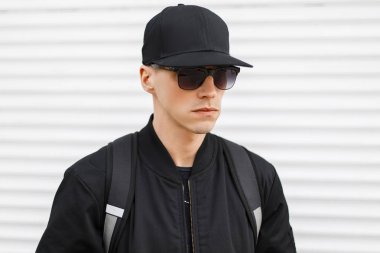 Bir moda siyah güneş gözlüğü metal beyaz duvarın yanında onun omuzlarında siyah bir sırt çantası ile trendy ceket siyah basketbol şapkalı şık çekici hipster adam. Amerikalı adam.