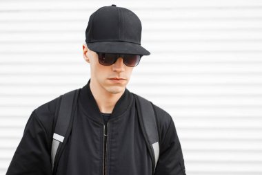 Şık siyah güneş gözlüğü onun omuzlarında beyaz bir arka plan üzerinde siyah bir sırt çantası ile şık bir ceket siyah şapkalı bir moda adam hipster portresi. Ciddi şık Amerikan adam.