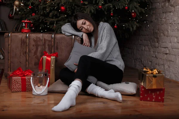 Genç kadın siyah pantolon beyaz çorap örme kazak sevimli bir gülümseme ile Noel ağacı Noel oda hediyeler arasında yakın katta oturuyor. Kız tatil hakkında düşünüyor.