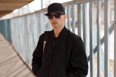 Genç çekici ciddi hipster adam ahşap bir çit arka plan yakınındaki bir siyah şapkalı şık siyah ceketli siyah güneş gözlüğü. Şık Amerikan adam.