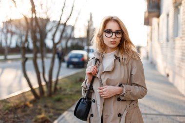 Genç hipster kadın moda deri çanta olan beyaz bir tişört trendy bardaklarda bahar paltolu şaşırtıcı bahar öğleden sonra sokakta yürüyor. Sevimli kız