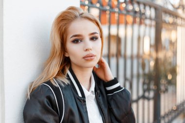 Bir metal geçit arka plan üzerinde beyaz bir duvar yakınındaki beyaz gömlekli bir şık siyah ceketli güzel gözlü çekici genç bir kadın portresi. Çekici kız.