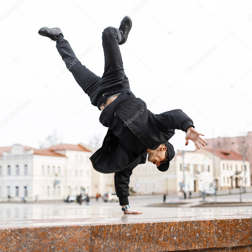 Stylish young man dancer in black jeans in a stylish jacket in a cap in sunglasses does a handstand in the city on the street. American hipster guy.