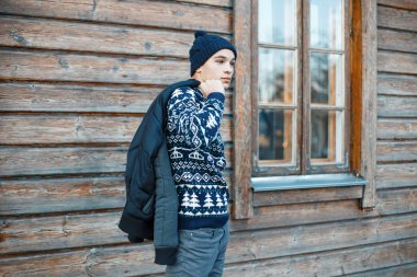 Ahşap kahverengi kır evi yakınındaki bir kış ceket konumu olan modaya uygun kot pantolon beyaz bir Noel desenli bir vintage kazak örme bir mavi şapkalı şık genç adam. Yakışıklı adam.