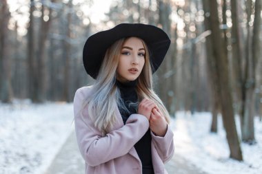 Şehvetli şık güzel genç kadın karlı bir ormandaki ağaçlar bir arka plan üzerinde Kış güneşli bir günde poz pembe zarif paltolu bir şık siyah şapkalı örgü vintage elbise giymiş. Güzel kız.