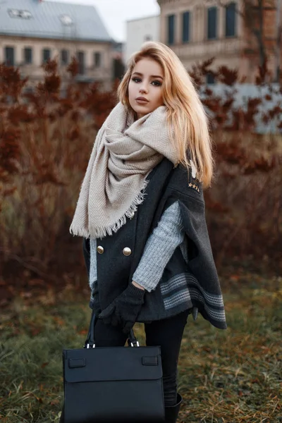 灰色でスタイリッシュなモダンな若い女性ニット レザー ファッション バッグと黒のジーンズにスカーフと黒いコートを着てセーター都市の乾いた草に近いスタンド 魅力的な女の子 — ストック写真