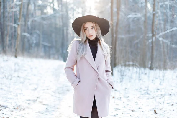 スタイリッシュな温かみのある華やかな服の非常に美しい若い女性は 冬の晴れた日に雪に覆われた森の中の優雅な帽子で歩いています ファッショナブルな魅力的なモダン ガール — ストック写真