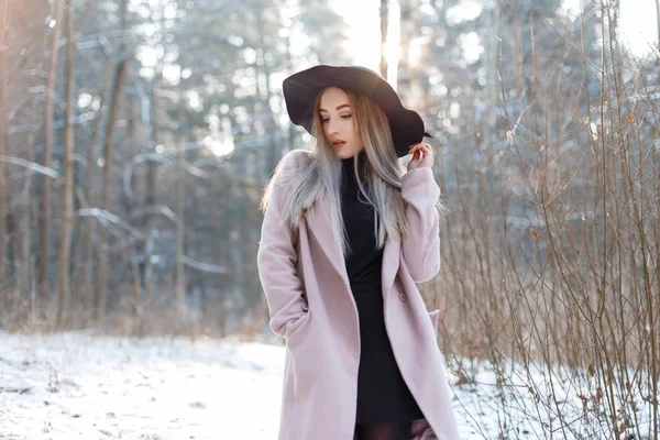 漂亮美丽的年轻女子穿着黑色针织礼服 在一个冬天的雪域公园里 穿着一顶优雅的黑色帽子 穿着一件时尚的粉色外套 在户外摆姿势 惊人的时尚金发女孩 — 图库照片
