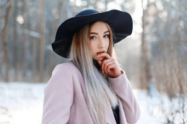 冬の森の背景にスタイリッシュな暖かいピンクのコートに黒のエレガントな帽子で魅力的な非常に若い女性の肖像画 美しいスタイリッシュな女の子 現代ファッション — ストック写真