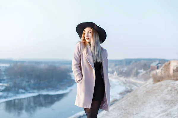 优雅时尚的年轻女子穿着粉色时尚外套 戴着别致的帽子 穿着黑色针织礼服 站在河的背景上的山上 时尚的女孩享受冬季景观 — 图库照片